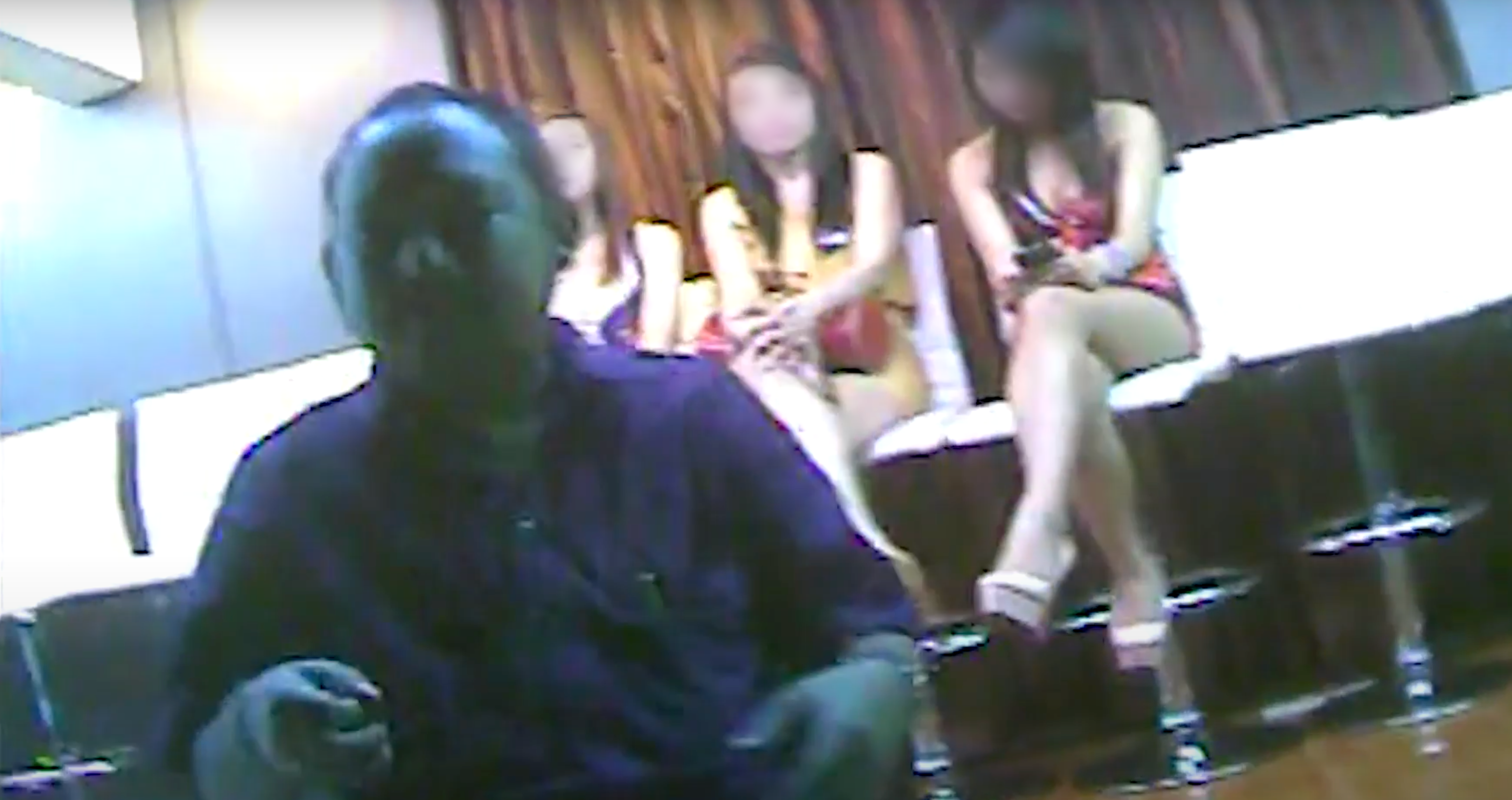 Three female victims of human trafficking sit, legs crossed, behind a male trafficker.